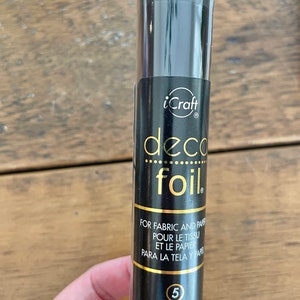 May include: A black tube of gold iCraft deco foil transfer sheets. The tube has a gold label with the text "deco foil" and "FOR FABRIC AND PAPER, POUR LE TISSU ET LE PAPIER, PARA LA TELA Y PAPEL". The label also includes the text "TRANSFER SHEETS, TRANSFERT FEUILLES, TRANSFERENCIA HOJAS" and "6 in x 12 in (15.2 cm x 30.5 cm)".