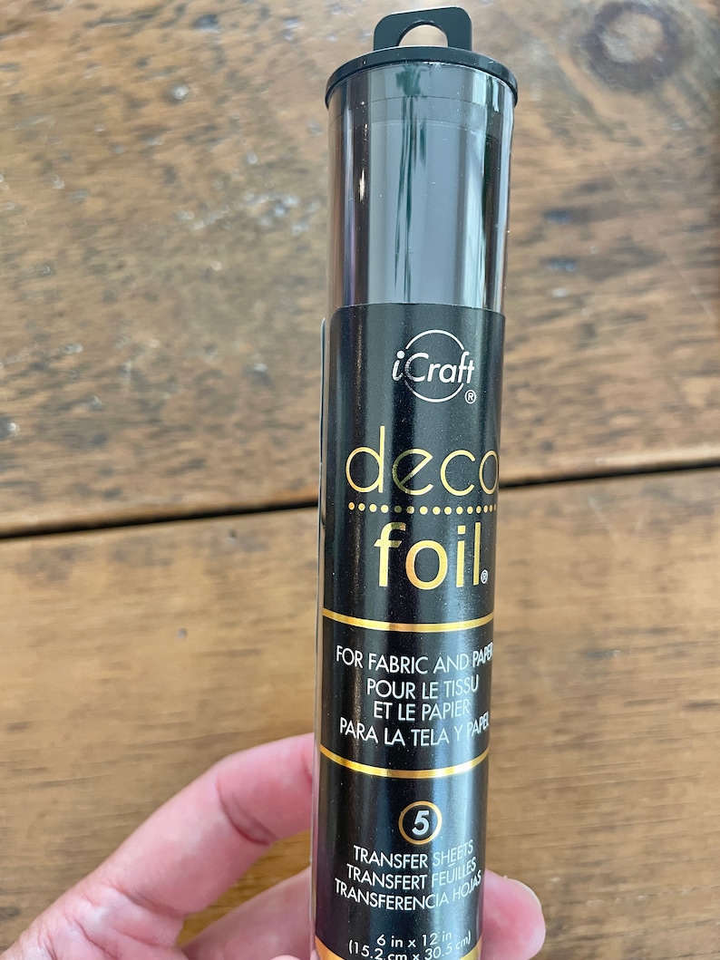 May include: A black tube of gold iCraft deco foil transfer sheets. The tube has a gold label with the text "deco foil" and "FOR FABRIC AND PAPER, POUR LE TISSU ET LE PAPIER, PARA LA TELA Y PAPEL". The label also includes the text "TRANSFER SHEETS, TRANSFERT FEUILLES, TRANSFERENCIA HOJAS" and "6 in x 12 in (15.2 cm x 30.5 cm)".