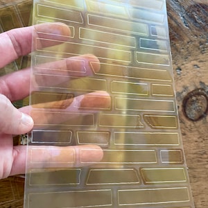 May include: A clear plastic sheet with a brick pattern stencil. The stencil is designed for use with paint or other crafting materials.