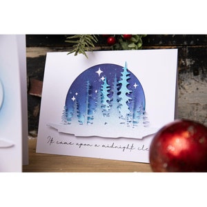 May include: A white card with a blue and white layered paper craft design of a snowy forest scene with the text "It came upon a midnight clear".