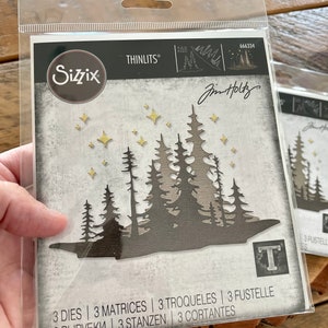 May include: A package of three metal dies for crafting, featuring a silhouette of a forest with stars. The dies are designed by Jim Holtz and are part of the Sizzix Thinlits collection. The package includes the product number 666334 and the text "3 Dies | 3 Matrices | 3 Troqueles | 3 Fustelle | 3 Вырубки | 3 Stanzen | 3 Cortantes".