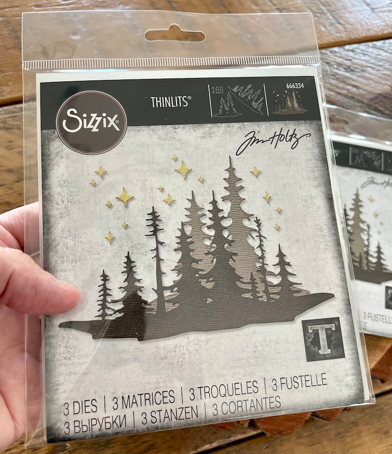 May include: A package of three metal dies for crafting, featuring a silhouette of a forest with stars. The dies are designed by Jim Holtz and are part of the Sizzix Thinlits collection. The package includes the product number 666334 and the text "3 Dies | 3 Matrices | 3 Troqueles | 3 Fustelle | 3 Вырубки | 3 Stanzen | 3 Cortantes".