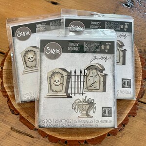 May include: A set of 22 metal dies for crafting, featuring a graveyard theme. The dies include a gate, tombstones, a skull, and a cross. The dies are packaged in a clear plastic bag with a white label that says "Sizzix Thinlits Colorize" and "Jim Holtz".
