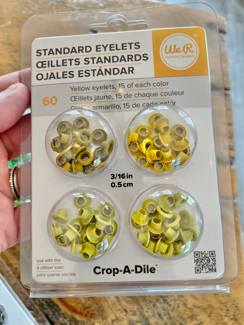 May include: A package of 60 yellow standard eyelets, 15 of each color. The eyelets are 3/16 inch (0.5 cm) in size and are packaged in clear plastic containers. The package is labeled "We R memory keepers" and "Crop-A-Dile".