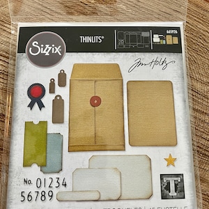 May include: A set of 45 die-cut metal craft dies featuring a vintage envelope, tags, and other decorative elements. The dies are designed by Jim Holtz and are part of the Sizzix Thinlits collection. The package includes the number 665926 and the text "No. 01234 56789".