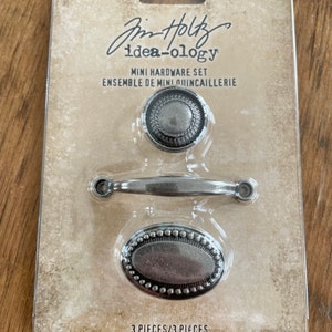 May include: A mini hardware set by Tim Holtz Idea-ology, featuring three antique silver-toned pieces: a round knob, a small handle, and an oval pull. The set is packaged on a cardboard backing with product information.