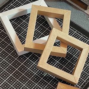May include: Three picture frames, one white and two brown, are stacked on top of each other. The frames are made of cardboard and have a simple design.