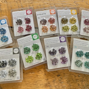 May include: Assortment of 10 packs of standard eyelets in various colors, including red, blue, green, purple, and gray. Each pack contains 60 eyelets and is labeled "Crop-A-Dile".
