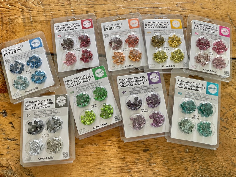 May include: Assortment of 10 packs of standard eyelets in various colors, including red, blue, green, purple, and gray. Each pack contains 60 eyelets and is labeled "Crop-A-Dile".