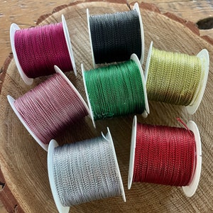 May include: Seven spools of colorful cord in shades of red, pink, green, silver, and black. The cord is tightly wound on white plastic spools. The spools are arranged on a wooden surface.