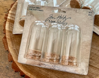 Peut inclure: Un paquet de trois dômes de présentation en verre transparent avec des bouchons en liège. L'emballage est étiqueté "Jim Holtz ide-ology" et "Display Domes/Dome d'Affichages". Les dômes sont étiquetés "Mini/Mini".