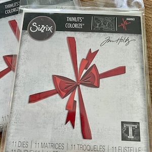 May include: A package of 11 red die-cut bow shapes for crafting. The package is labeled "Sizzix Thinlits Colorize" and "Jim Holtz". The package includes the text "11 Dies | 11 Matrices | 11 Troqueles | 11 Fustelle | 11 Вырубки | 11 Stanzen | 11 Cortantes".