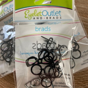 May include: A clear plastic bag containing a variety of black and silver metal eyelets. The bag has a green and white label that reads "Eyelet Outlet and Brads" and "brads". The label also includes the text "Ages 8+".