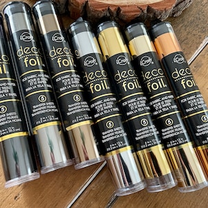 May include: A set of eight metallic transfer sheets for crafting. The sheets are packaged in clear tubes with black labels that read "iCraft deco foil". The labels also include the text "For fabric and paper, pour le tissu et le papier, para la tela y papel" and "5 transfer sheets, transfert feuilles, transferencia hojas". The sheets are available in silver, gold, copper, and black.
