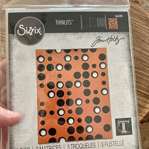 May include: A Sizzix Thinlits die set featuring a copper-colored die with a pattern of black and white circles. The package includes the text "Sizzix", "Thinlits", and "Jim Holtz". The package also states "3 DIES | 3 MATRICES | 3 TROQUELES | 3 FUSTELLE".