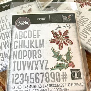 May include: A set of 43 metal dies for crafting, featuring letters, numbers, and holiday-themed designs. The dies are silver with a textured finish and include a poinsettia, holly, and a letter T. The packaging includes the text "Sizzix Thinlits" and "43 Dies | 43 Matrices | 43 Troqueles | 43 Fustelle | 43 Вырубки | 43 Stanzen | 43 Cortantes".