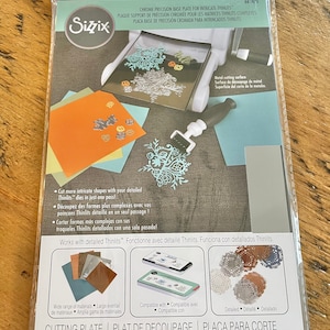 May include: Sizzix Chrome Precision Base Plate for intricate Thinlits. The package shows a metal cutting surface and examples of cut shapes. It includes the text "Cut more intricate shapes with your detailed Thinlits dies in just one pass!"