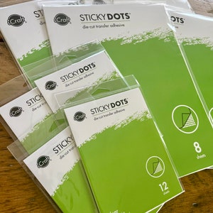 May include: A stack of clear adhesive sheets in plastic packaging. The packaging is green and white with the text "STICKY DOTS die-cut transfer adhesive". The sheets are available in packs of 8 and 12. The sheets are 4.25 inches by 5.5 inches and are acid free and permanent.