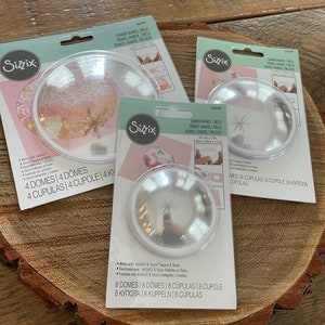 May include: Three clear plastic shaker domes for crafting projects. The domes are packaged on white cards with the Sizzix logo and product information. The domes are available in three sizes: 4, 6, and 8 inches in diameter.
