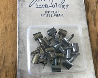 May include: A package of 15 small, antique gold-toned metal clips. The package is labeled "Tim Holtz idea-ology Tiny Clips Petits L'agrafe 15 Clips/15 Agrafes".