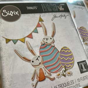 May include: Sizzix Thinlits die set featuring Easter-themed designs. The set includes bunny figures, decorated Easter eggs, and a banner. The packaging displays the "Jim Holtz" signature and the Thinlits logo. The die cuts are suitable for paper crafting.