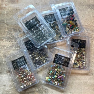 May include: Six clear plastic packages of 100 paper fasteners. Each package contains a different color of fasteners, including silver, gold, black, red, blue, and green. The packages have the text "100 Paper Fasteners" and "Create!" printed on them.