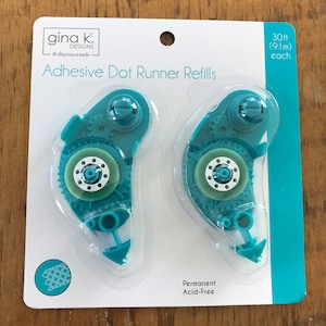 May include: Two turquoise blue adhesive dot runner refills, each with a clear plastic cover and a white gear mechanism. The packaging is white with black text that reads "gina k. DESIGNS thermo web Adhesive Dot Runner Refills 30ft (9.1m) each Permanent Acid-Free".