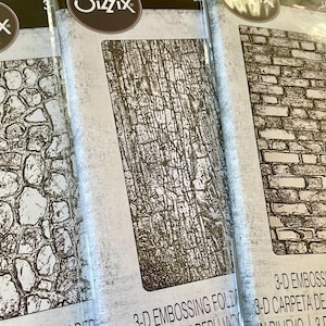 May include: Three Sizzix 3-D embossing folders. The folders feature stone, bark, and brick patterns. The packaging includes the Sizzix logo and product information. These folders are designed for crafting and paper projects.
