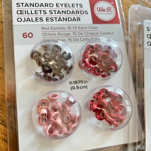 May include: A package of 60 standard eyelets in three colors: red, brown, and pink. The eyelets are 0.1875 inches (0.5 cm) in diameter. The package is labeled "We R Memory Keepers".