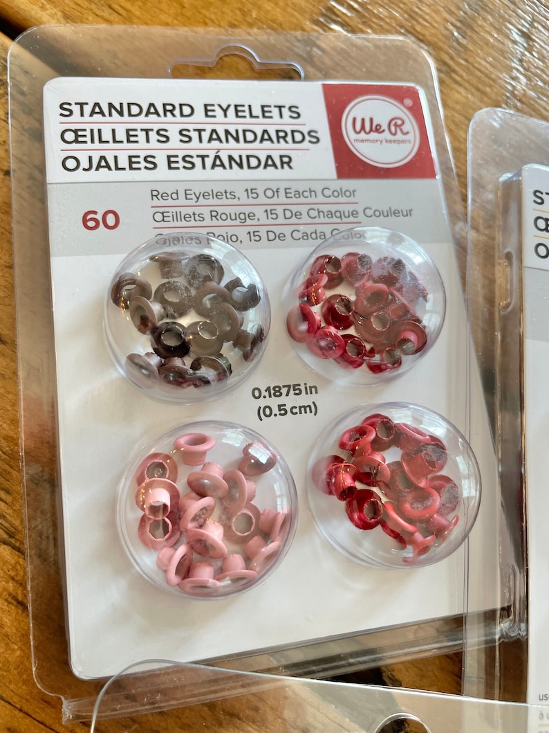 May include: A package of 60 standard eyelets in three colors: red, brown, and pink. The eyelets are 0.1875 inches (0.5 cm) in diameter. The package is labeled "We R Memory Keepers".