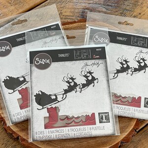 May include: Three packages of Sizzix Thinlits dies featuring Christmas-themed designs. Each package displays a black silhouette of Santa's sleigh and reindeer, along with a chimney and the letter "T". The packaging includes the text "Jim Holtz".