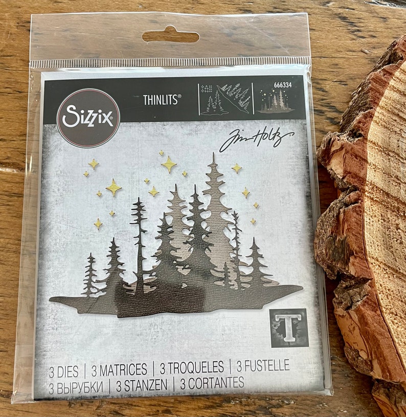 May include: A package of three metal dies for a craft cutting machine. The dies are designed to cut out a silhouette of a forest with trees and stars. The package is labeled "Sizzix Thinlits" and "Jim Holtz".