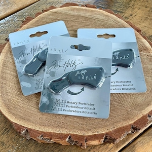 May include: Three packages of a black mini rotary perforator by Jim Holtz Tonic Studios. The perforator is designed to create a curved cut. The packaging features the Jim Holtz Tonic Studios logo and the text "Mini Rotary Perforator", "Mini Perforateur Rotatif", and "Mini Perforadora Rotatoria".