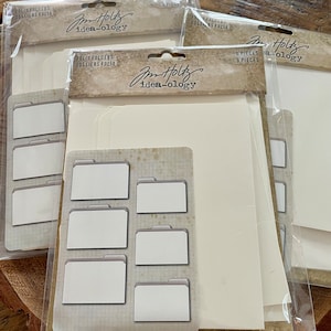 May include: A set of six white folder dividers with a grid pattern and the text "Tim Holtz idea-ology" on the packaging. The dividers are designed for organizing and labeling projects.