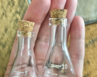 Peut inclure: Deux petites fioles en verre transparent avec des bouchons en liège. Les fioles ont une forme conique avec un fond plat et des marquages de mesure blancs. Une fiole a des marquages pour 3 et 5, l'autre pour 5 et 10.