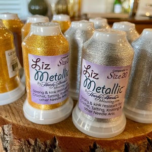 May include: Close-up of several spools of Liz Metallic thread in gold and silver. The spools have white bases and tops, with labels that read "Liz Metallic" and "Size 20". The thread is designed for crochet, tatting, knitting, and other needle arts.