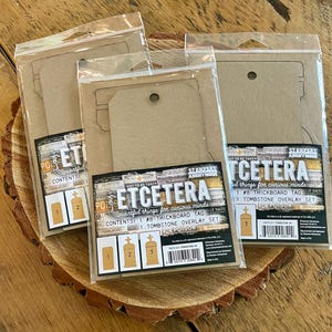 May include: Four sealed packages of craft supplies on a wooden surface. Each package contains a thickboard tag and a tombstone overlay set. The packages have the text "ETCETERA" and "POISON" printed on them.