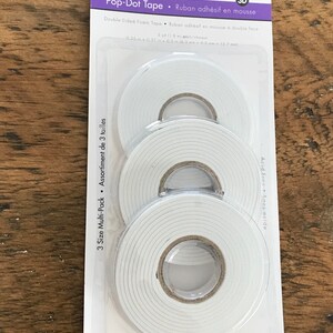 May include: A package of three rolls of double-sided foam tape. The tape is white and comes in three different sizes. The package is labeled "Pop-Dot Tape" and "Ruban adh&eacute;sif en mousse".