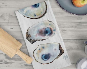 May include: A white kitchen towel with three watercolor illustrations of oyster shells. The shells are painted in shades of blue, brown, and white. The towel has a small logo in the bottom right corner that says "The Painted Oyster".