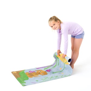 Puede incluir: Una esterilla de yoga rectangular con fondo azul claro que presenta una ilustración de dibujos animados de un perro marrón, hierba verde y flores moradas. La esterilla está parcialmente enrollada, revelando una superficie lisa con un diseño gráfico colorido y divertido.