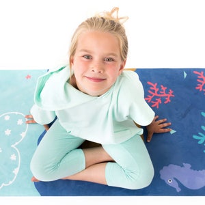 Puede incluir: Una alfombra de juego rectangular con un diseño de dos tonos, que presenta una sección azul claro con patrones de nubes y estrellas blancas y una sección azul oscuro decorada con corales, formas geométricas y un caballito de mar morado.