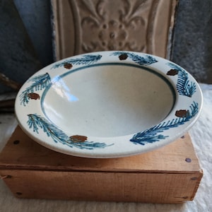 May include: A white ceramic bowl with a blue and gold pine needle design around the rim. The bowl is sitting on a wooden box.
