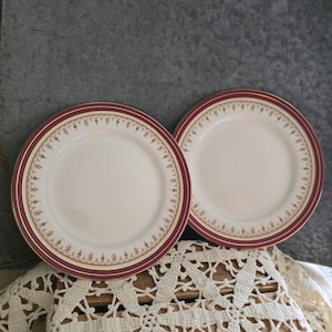 May include: Two white ceramic plates with a burgundy border and gold trim. The plates have a decorative gold pattern around the rim. The plates are displayed on a wooden surface with a white lace doily.