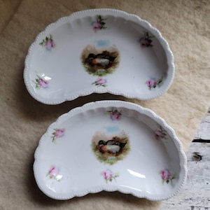 May include: Two white porcelain half-moon shaped serving dishes with a scalloped edge. Each dish features a painted image of a duck in a pond with pink roses around the edge.