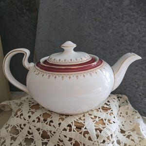 May include: White porcelain teapot with a rounded body, a curved handle, and a spout. The lid and a band around the middle are burgundy with gold trim and detailing. The teapot is resting on a white lace doily.