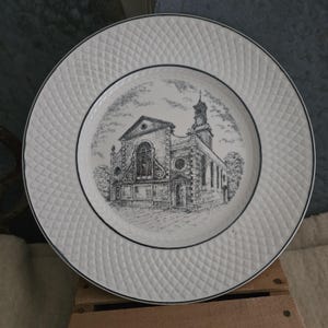 May include: A round white ceramic plate featuring a black and white sketch of a church building in the center. The rim of the plate has a textured diamond pattern and a thin black circular border line that frames the central illustration.