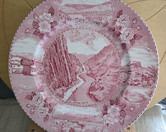 May include: A round ceramic plate featuring a maroon and white transferware design of Rocky Mountain National Park. The centre depicts Big Thompson Canyon, surrounded by four vignettes of Estes Park, Trail Ridge Road, Bear Lake, and Hallett Peak.