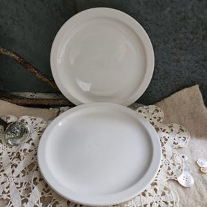 May include: Two round, off-white ceramic plates with a smooth, glossy finish and a wide, flat rim. The plates are stacked vertically, featuring a simple, clean design with no patterns or decorations, showcasing a uniform, neutral color throughout.