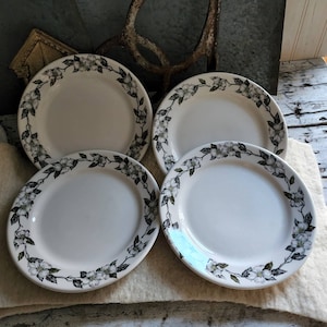 May include: Four white ceramic plates with a floral border design. The border features white flowers and green leaves against a black background. The plates are arranged on a light-colored surface.