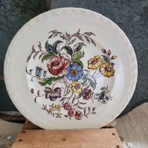 May include: Cream-colored ceramic plate with a floral design. The plate features a central bouquet of flowers in shades of red, blue, yellow, and purple, with green leaves. The rim has a textured, rope-like border. A vintage-style decorative plate.